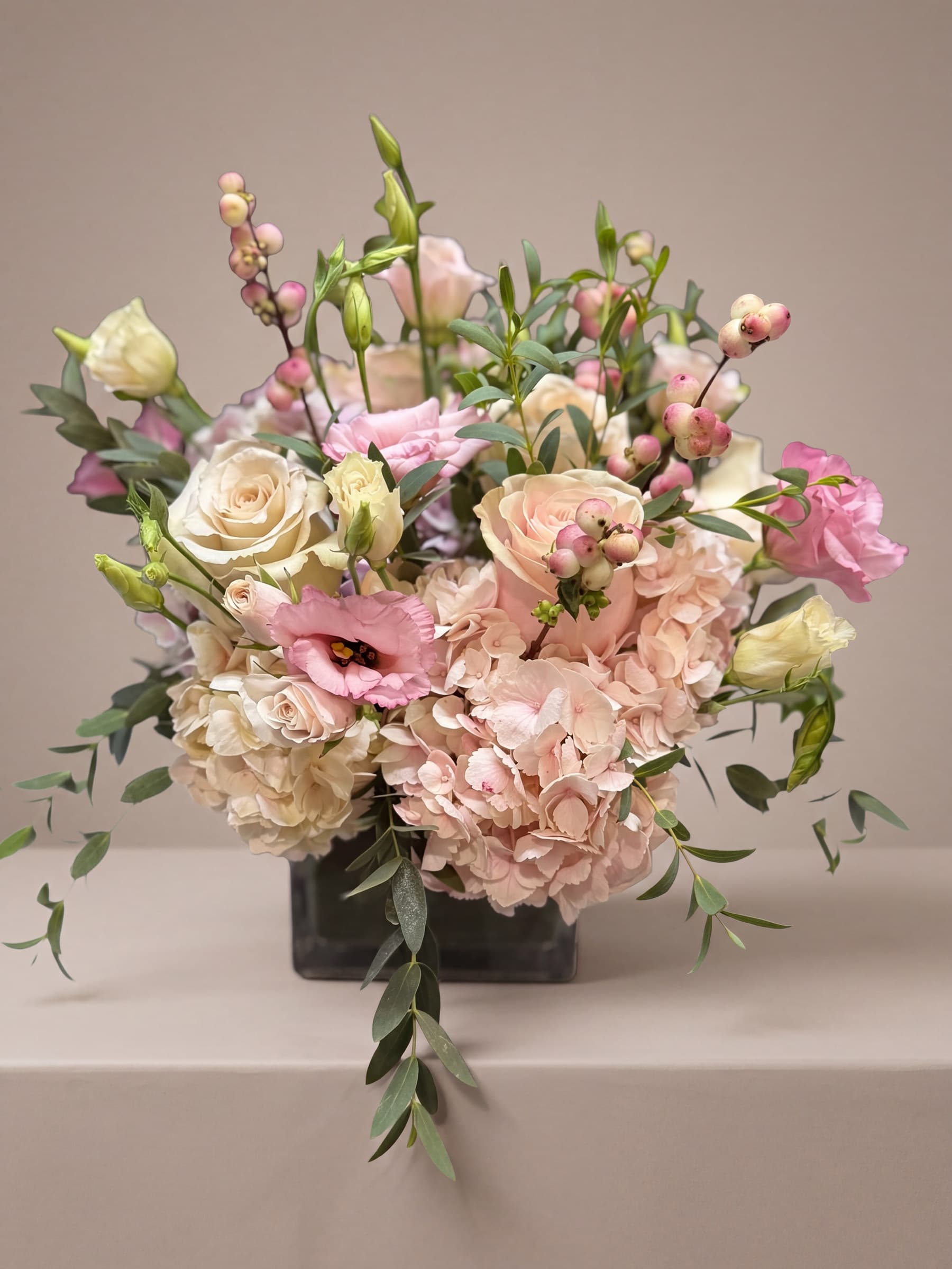 Low blush arrangement of pale pink hydrangea, cream roses, pink lisianthus, snowberry and trailing eucalyptus in a glass cube