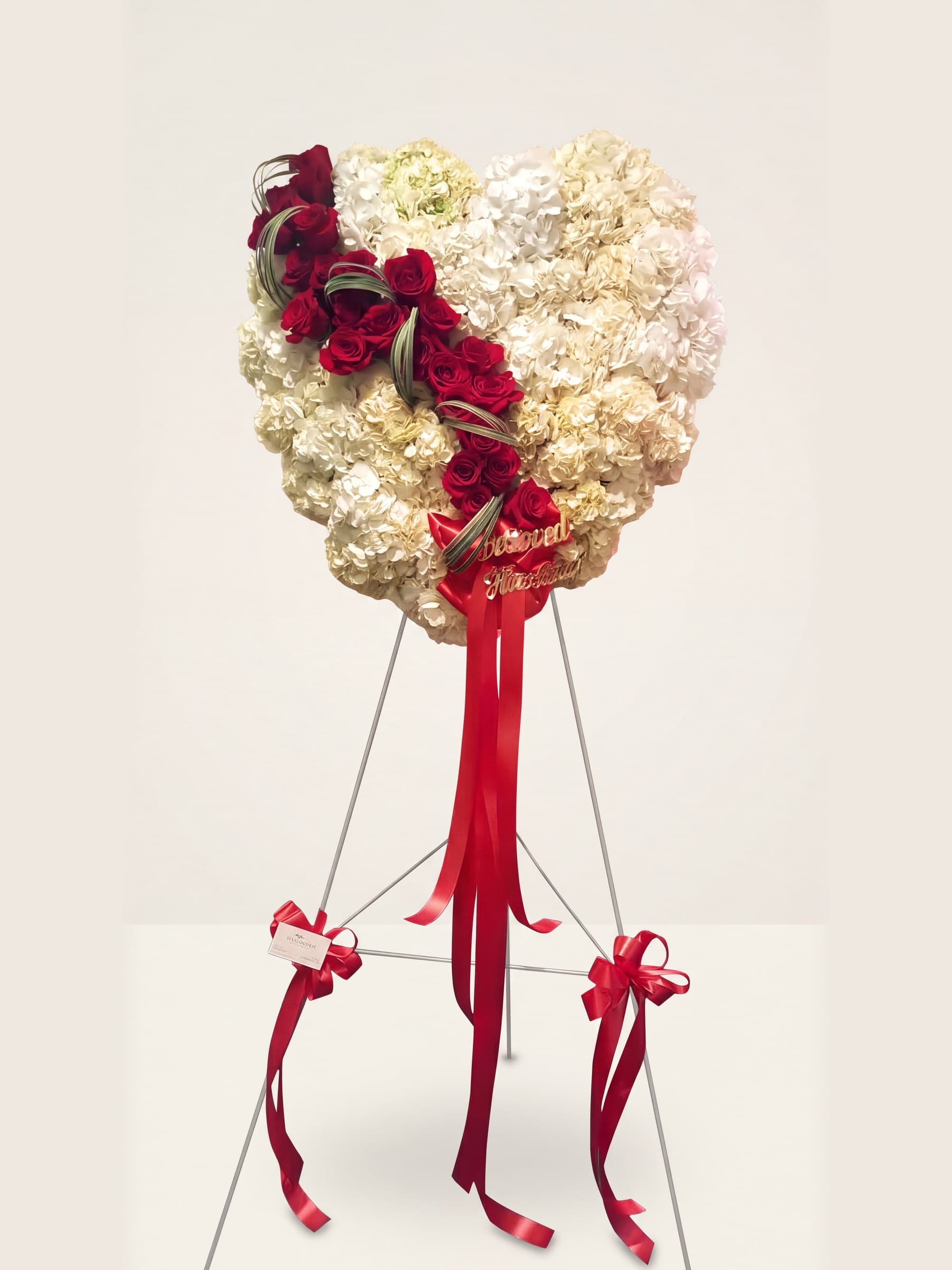 Funeral heart of white hydrangea with a diagonal cascade of red roses and red ribbons on an easel
