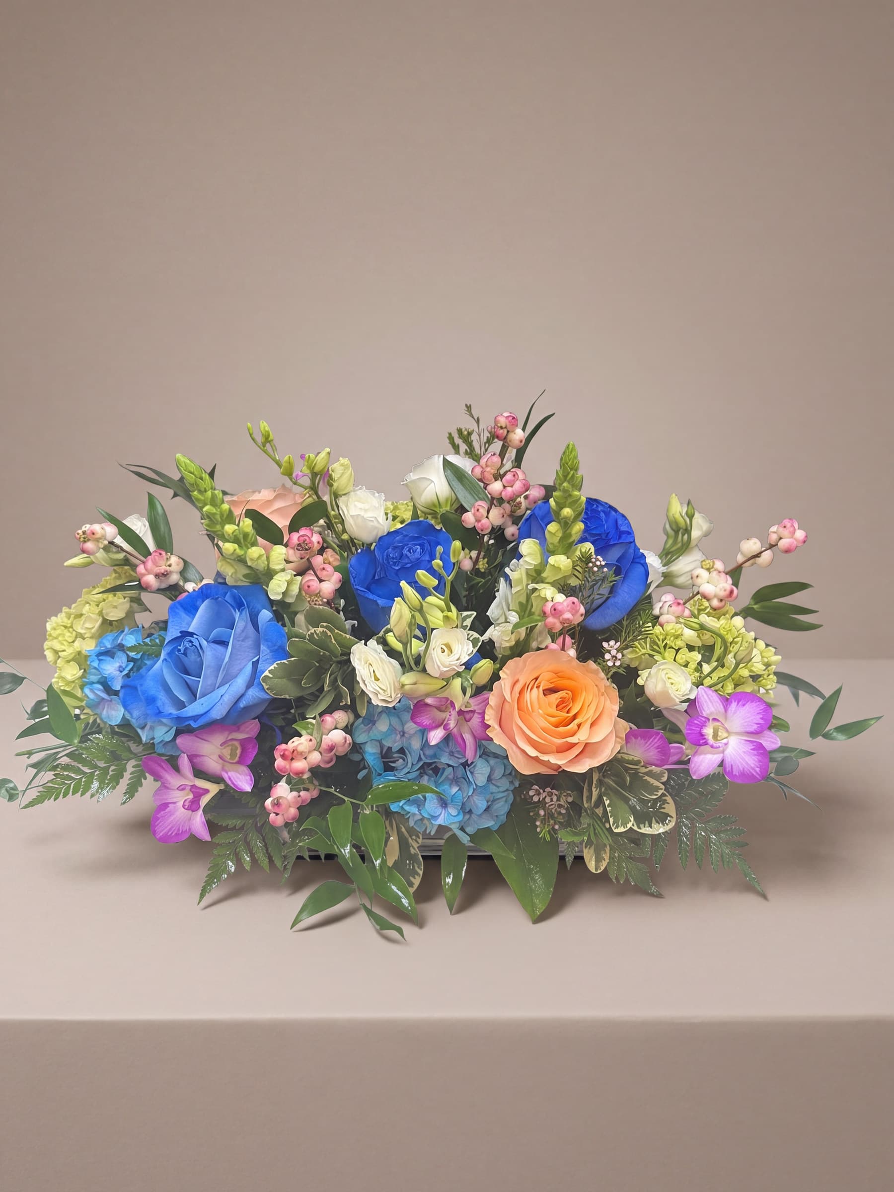 Low oval centerpiece of blue-tinted and peach roses, purple dendrobium orchids, blue and green hydrangea, white lisianthus and snapdragons over fern and ruscus