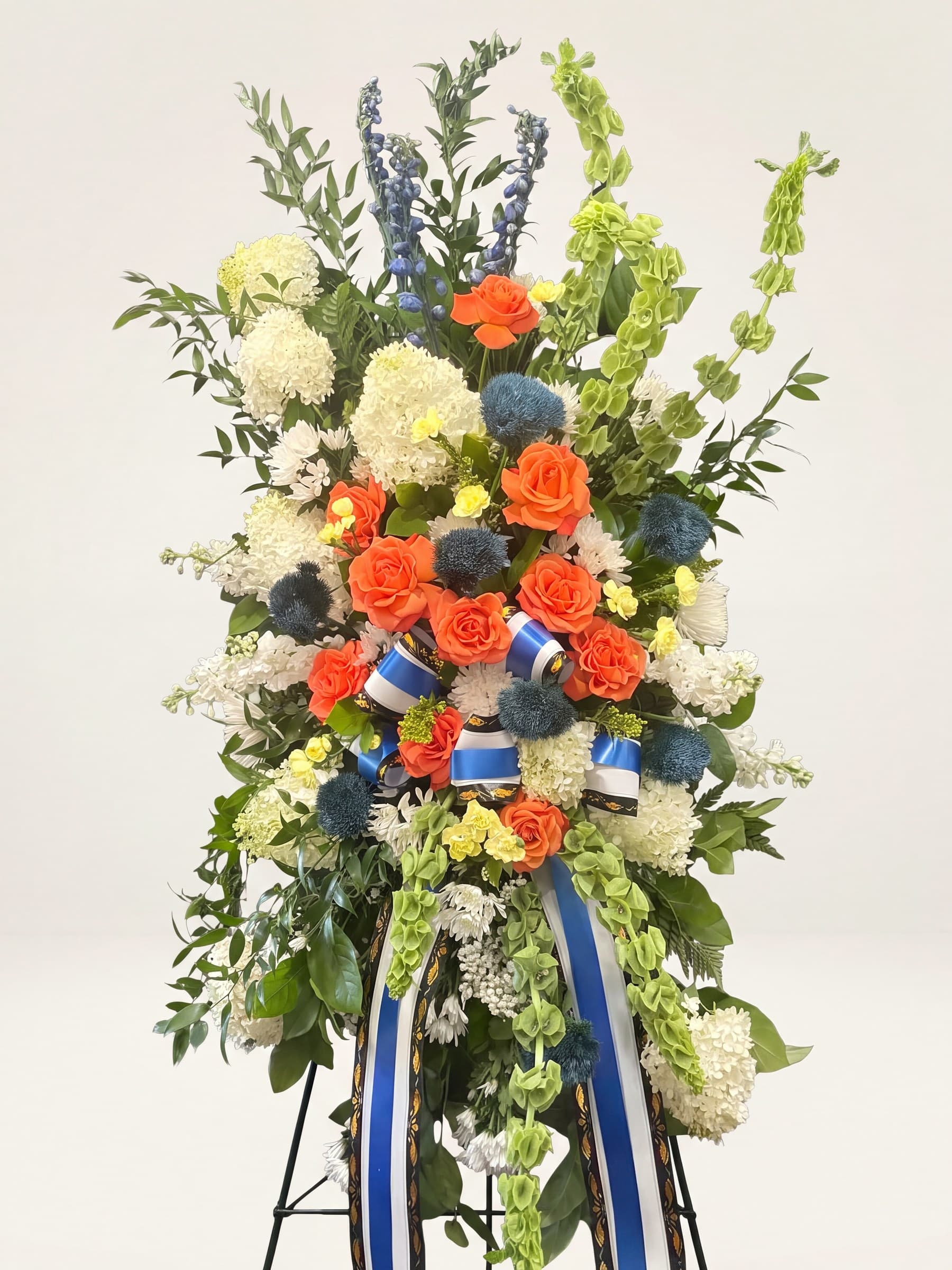 Standing funeral spray of white hydrangea and orange roses with blue thistle, delphinium, and a blue-and-white ribbon