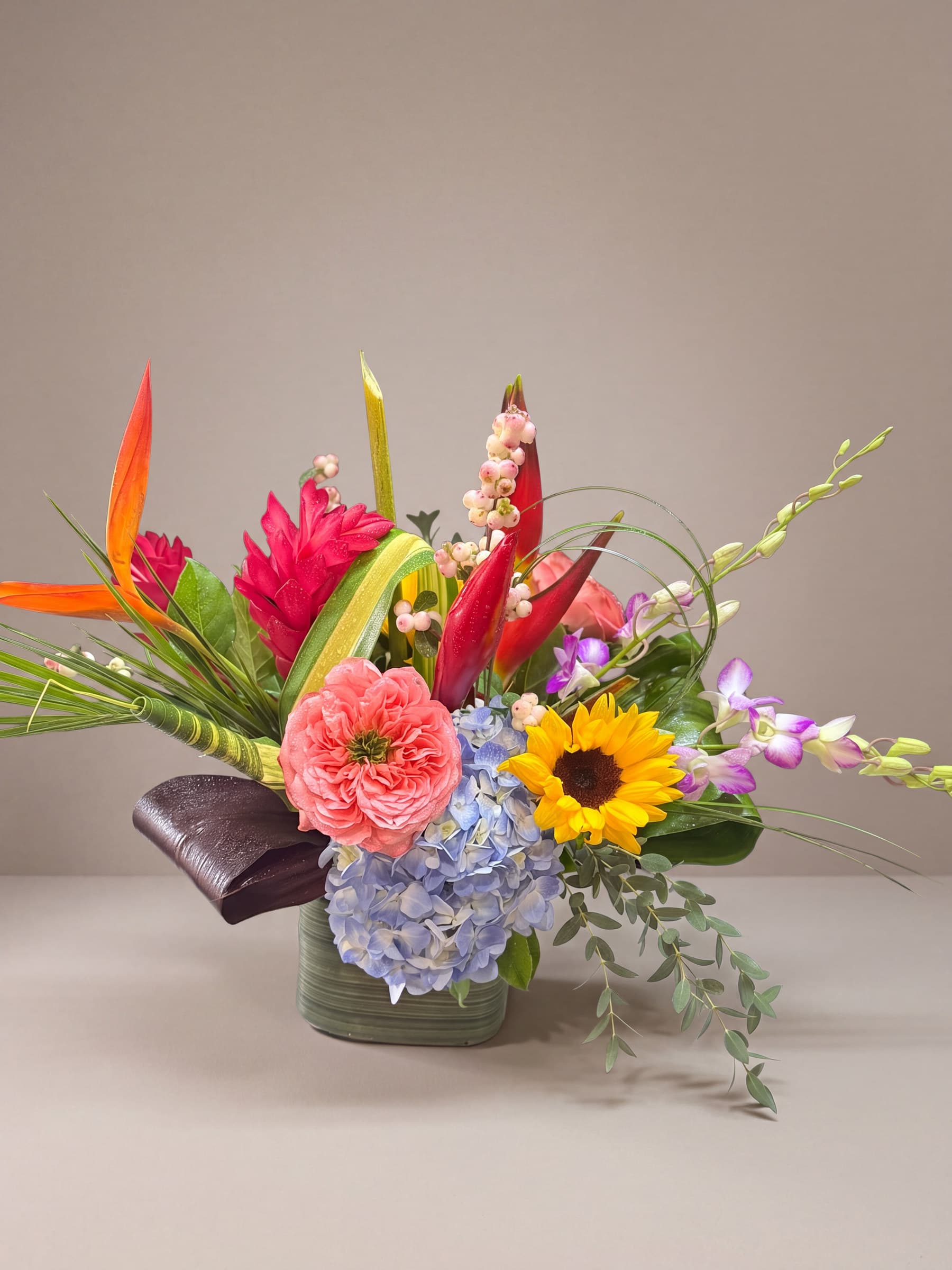 Sculptural tropical arrangement of bird of paradise, red heliconia, pink ginger, sunflower, blue hydrangea, coral garden rose and purple dendrobium orchids in a leaf-lined glass cube