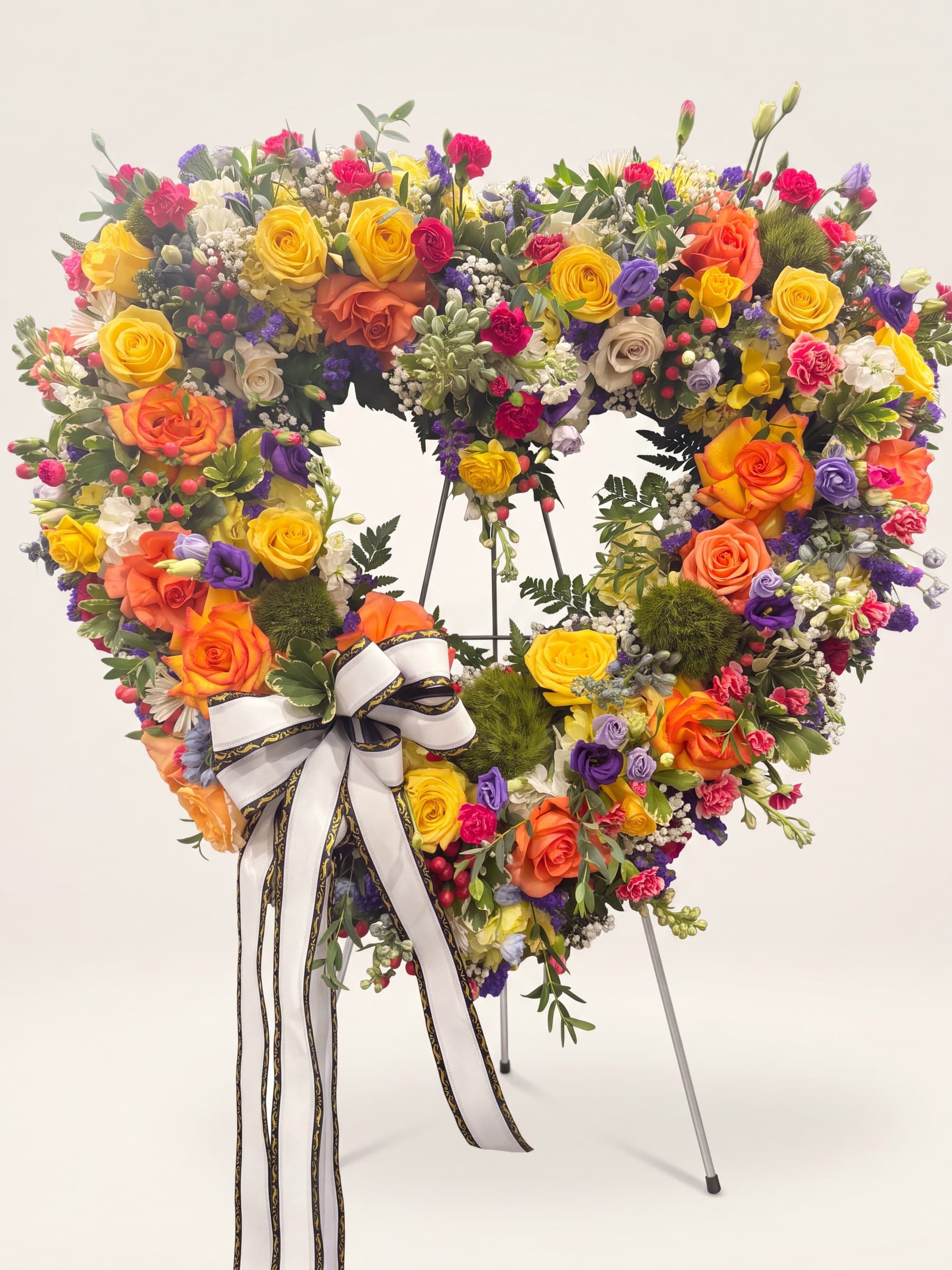 Open-heart funeral wreath of yellow and orange roses, carnations, and purple lisianthus with a white-and-black bow on an easel