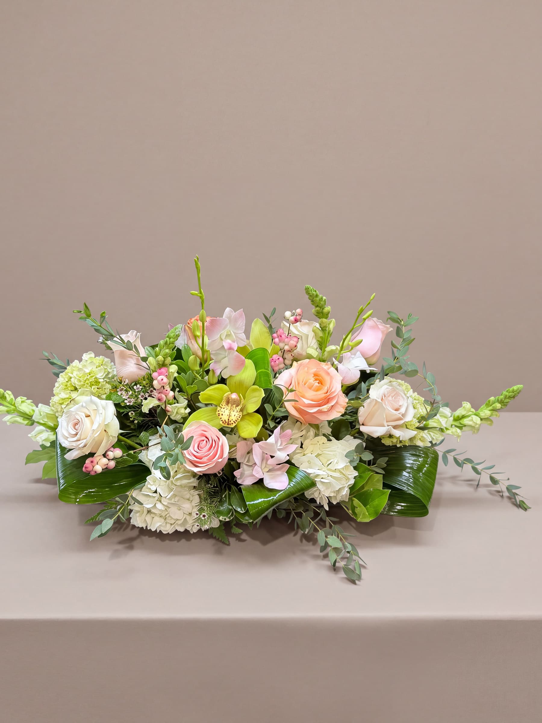 Long low oval centerpiece of peach and blush roses, green cymbidium and white dendrobium orchids, white hydrangea and snapdragons over aspidistra leaves and eucalyptus