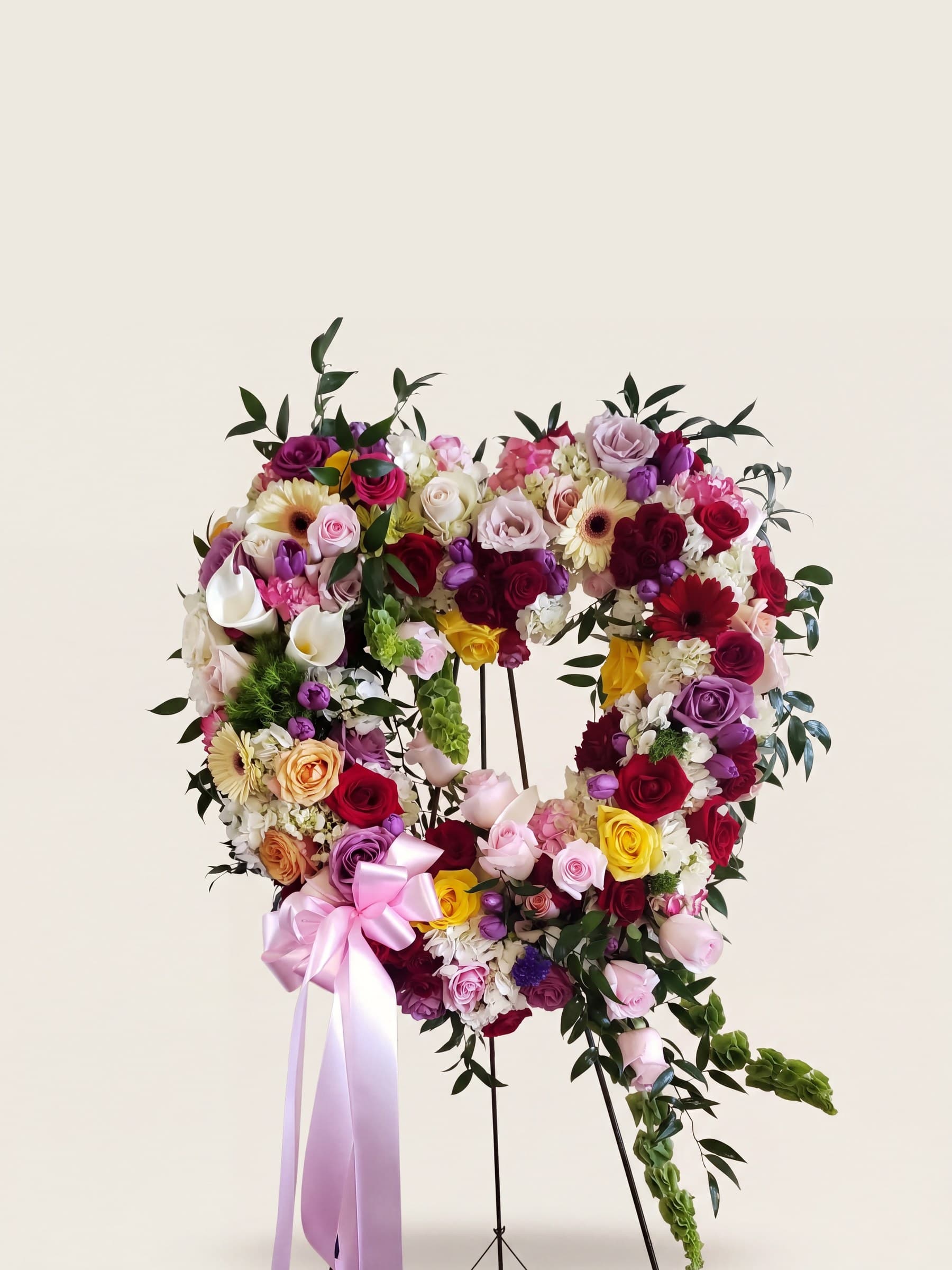 Open-heart funeral wreath of roses and gerbera daisies in many colors with a pink ribbon on an easel