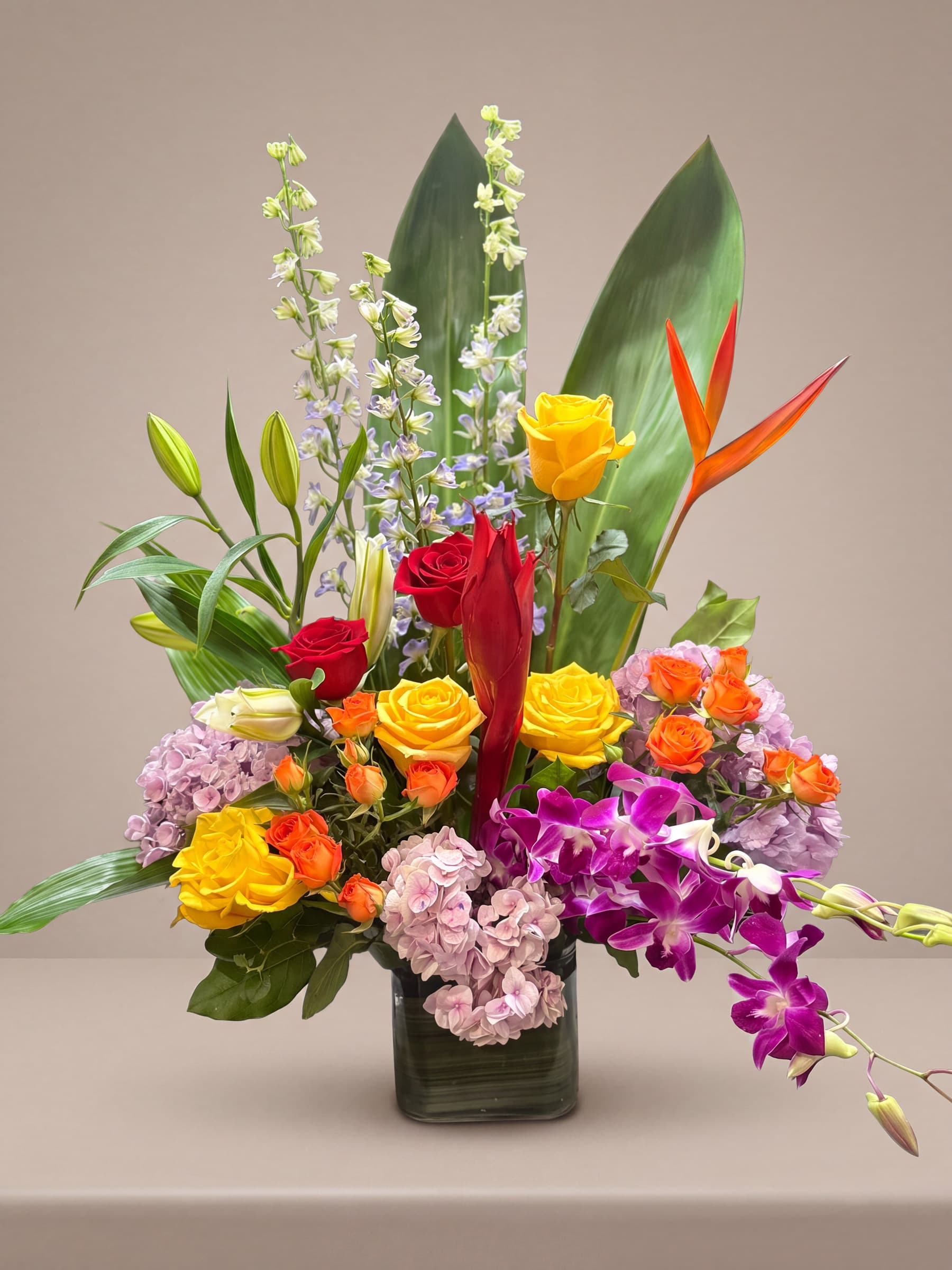 Tall tropical arrangement of orange heliconia, red ginger, yellow and red roses, lavender hydrangea and purple dendrobium orchids in a leaf-lined glass cube