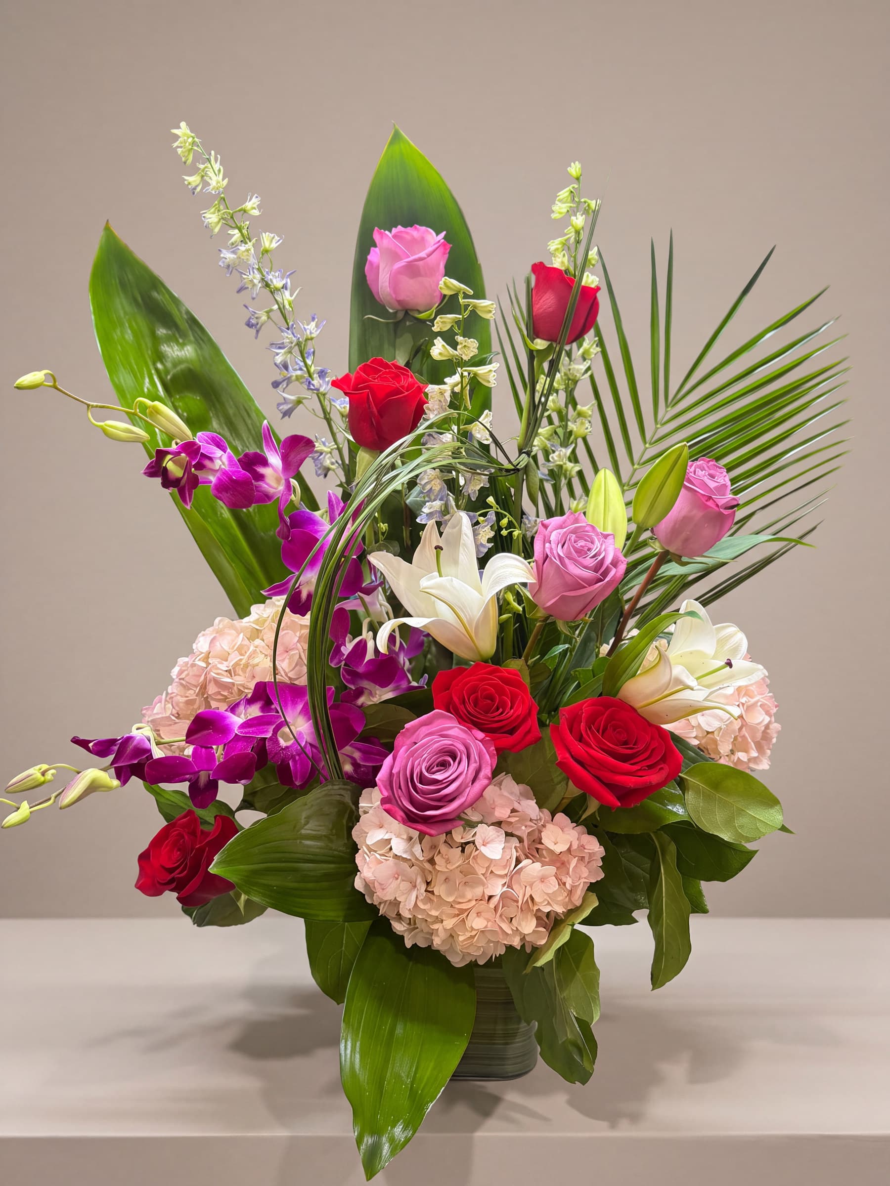 Tall arrangement of lavender and red roses, purple dendrobium orchids, white lilies and blush hydrangea with tropical leaves in a leaf-lined glass vase