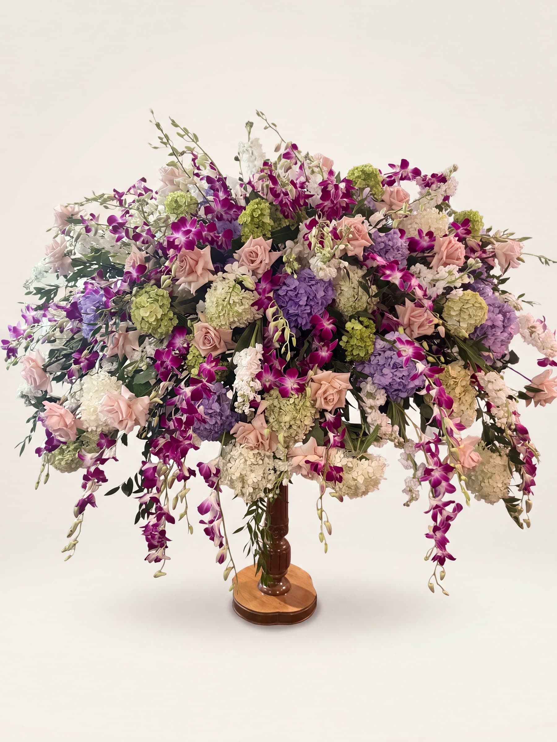 Grand funeral arrangement on a wooden pedestal with cascading fuchsia orchids, lavender hydrangea, and blush roses
