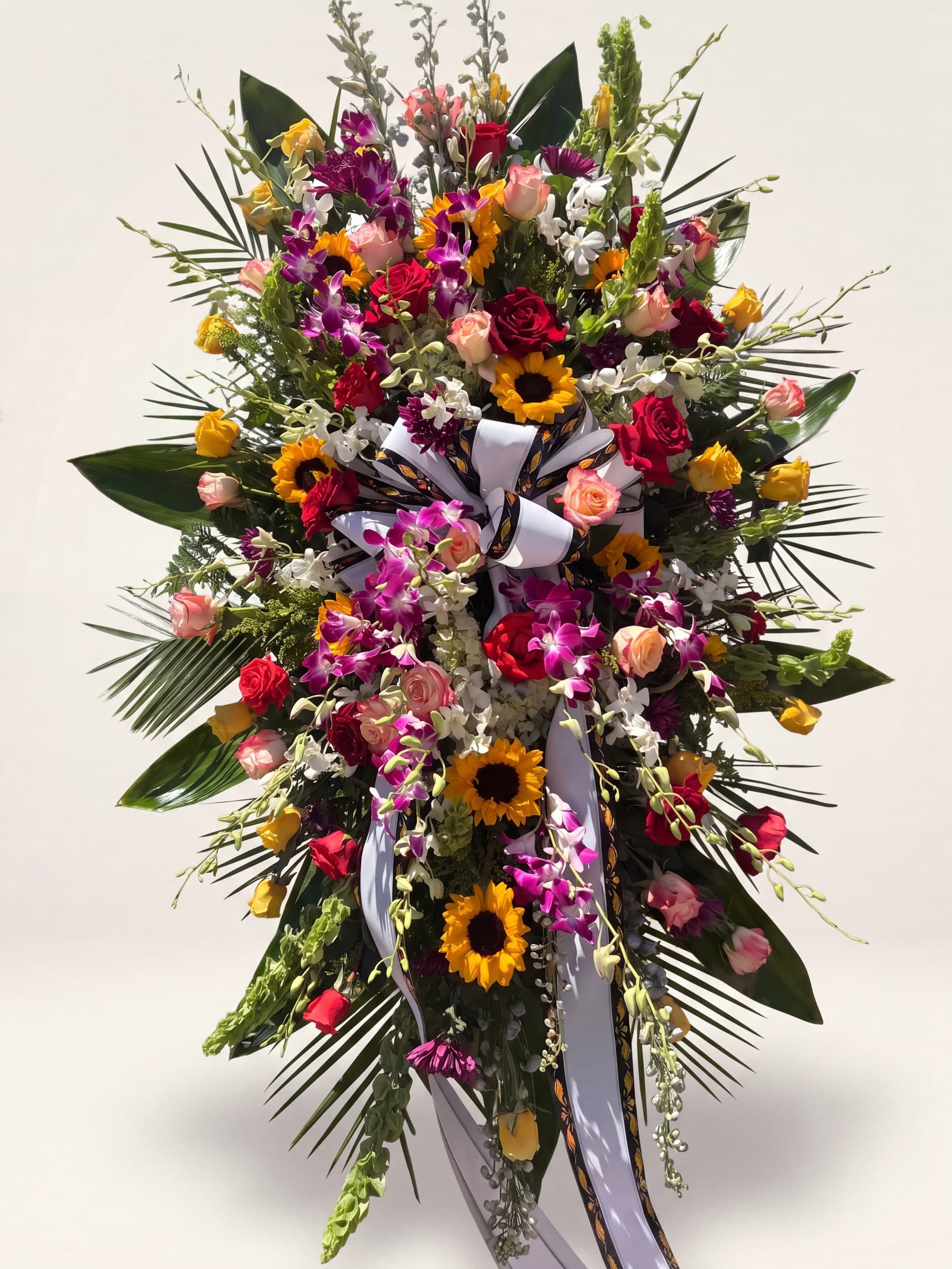 Large standing funeral spray of sunflowers, fuchsia orchids, and red, peach, and yellow roses with a black-and-white bow