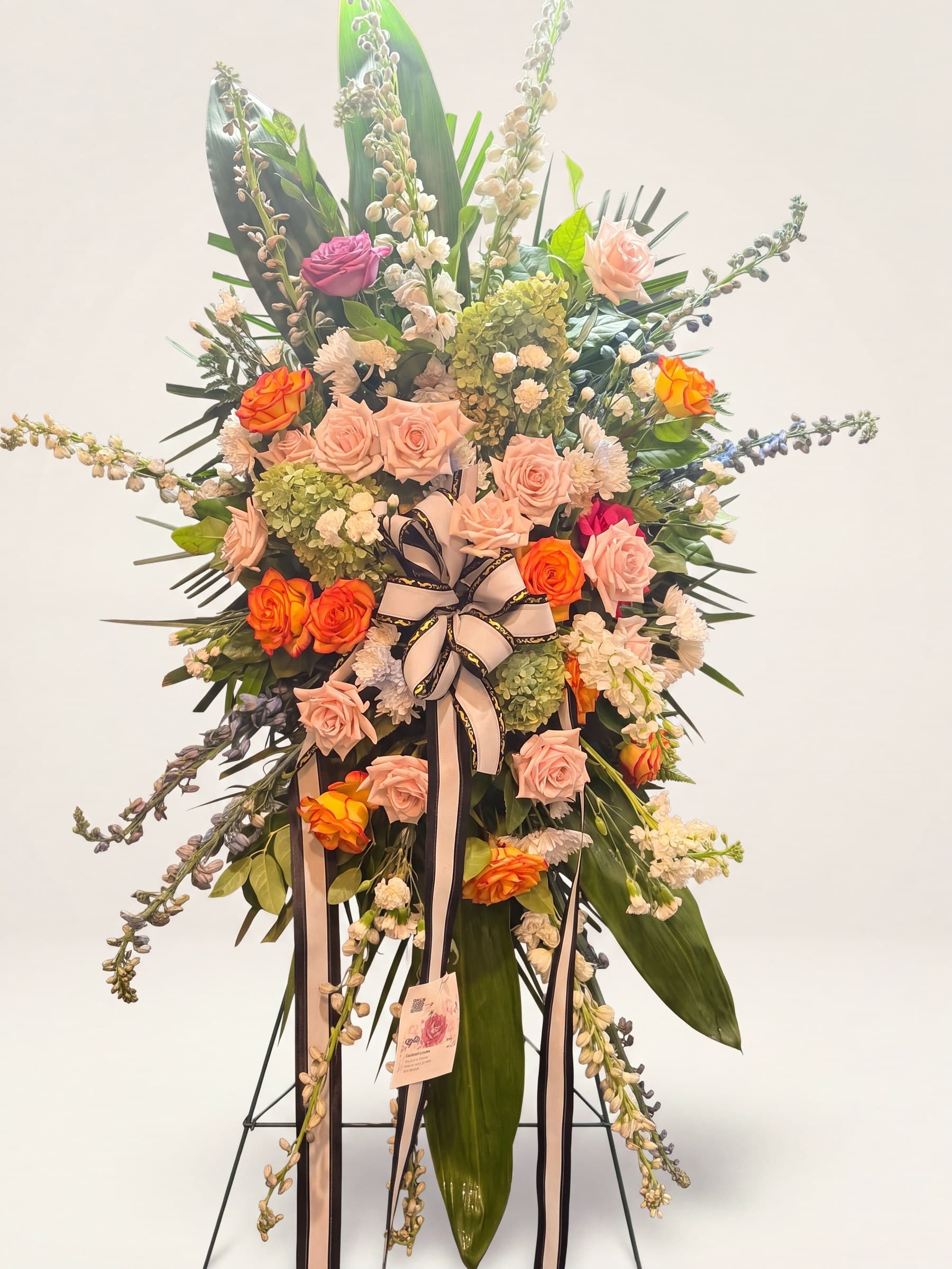 Standing funeral spray of peach and orange roses with a black-and-white bow on an easel