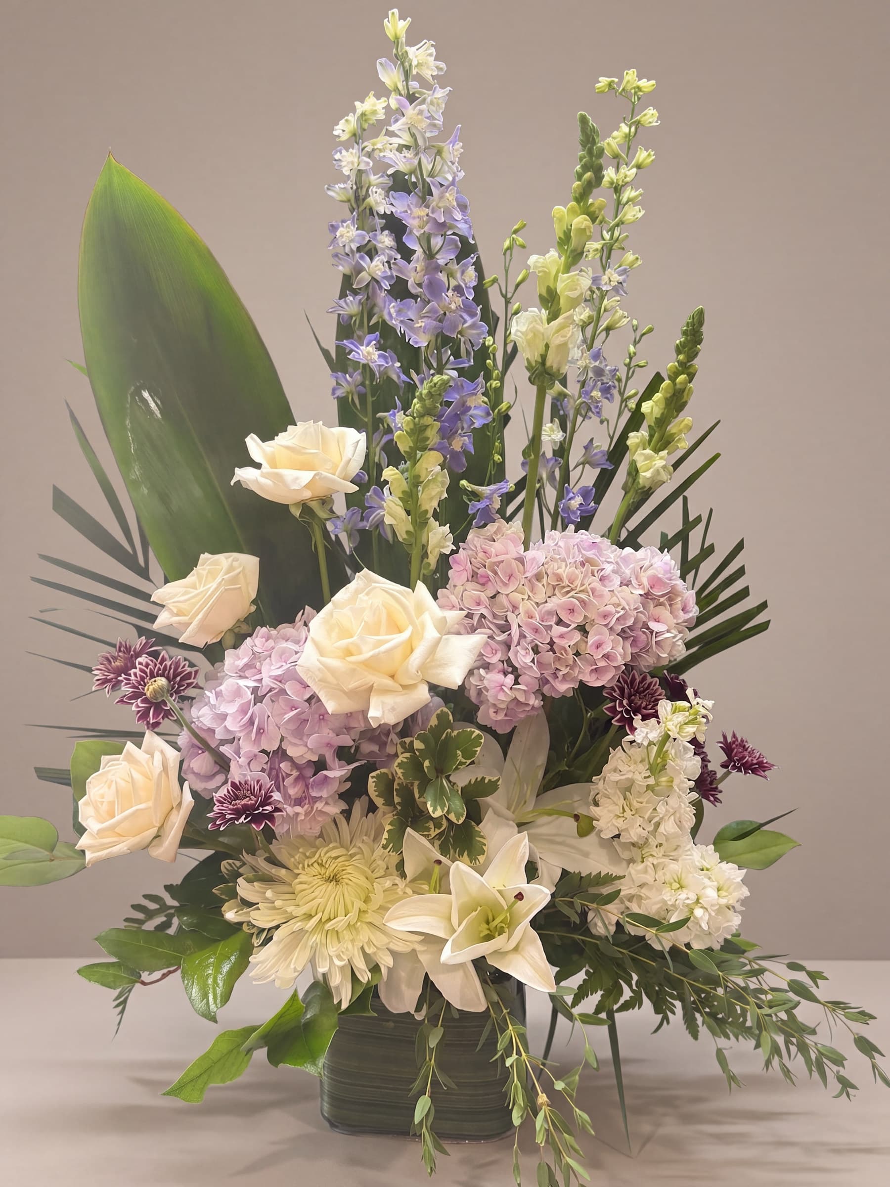 Standing sympathy tribute of white roses, lavender hydrangea, blue delphinium, white lilies, snapdragon and stock with burgundy chrysanthemum buttons