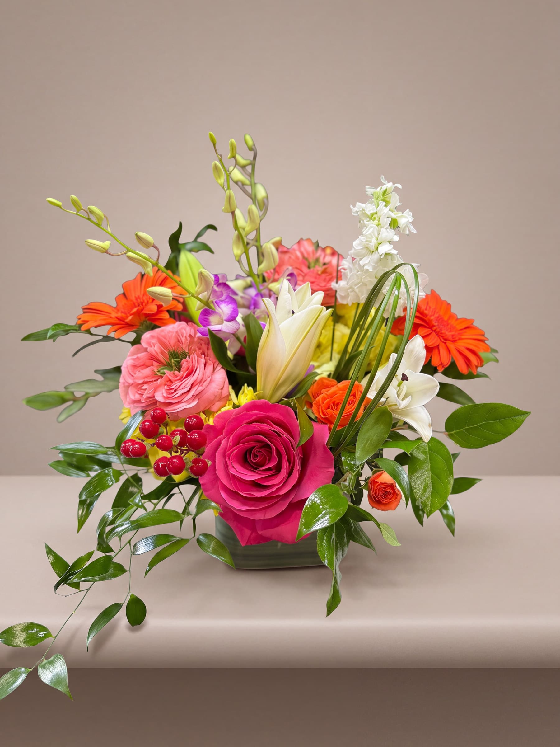 Bright arrangement of hot pink and coral garden roses, orange gerbera, purple dendrobium orchids, white lilies and stock with red berries in a leaf-lined glass cube