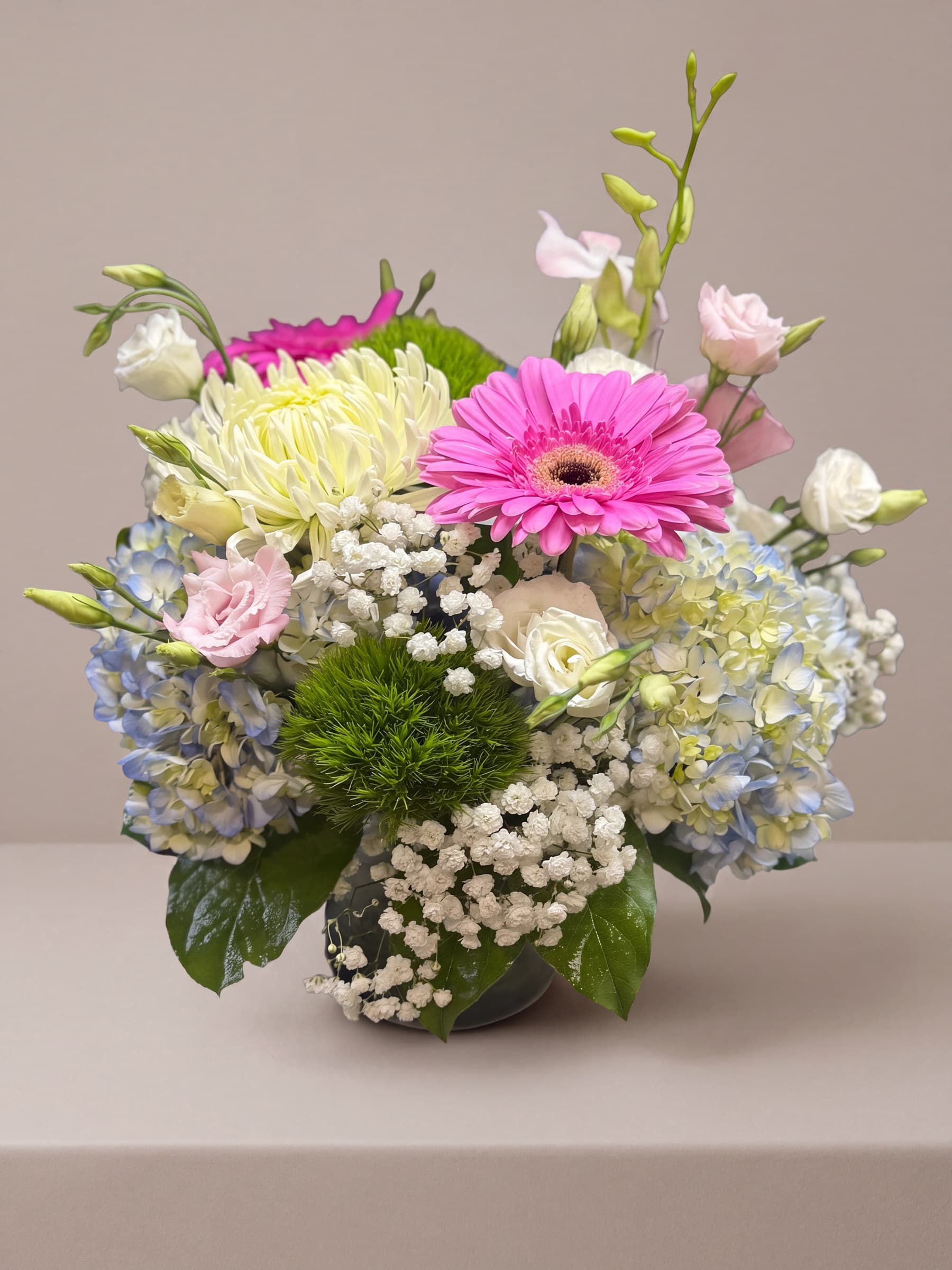 Low round arrangement of blue hydrangea, pink gerbera, white football mums, lisianthus and baby's breath in a leaf-lined glass cube