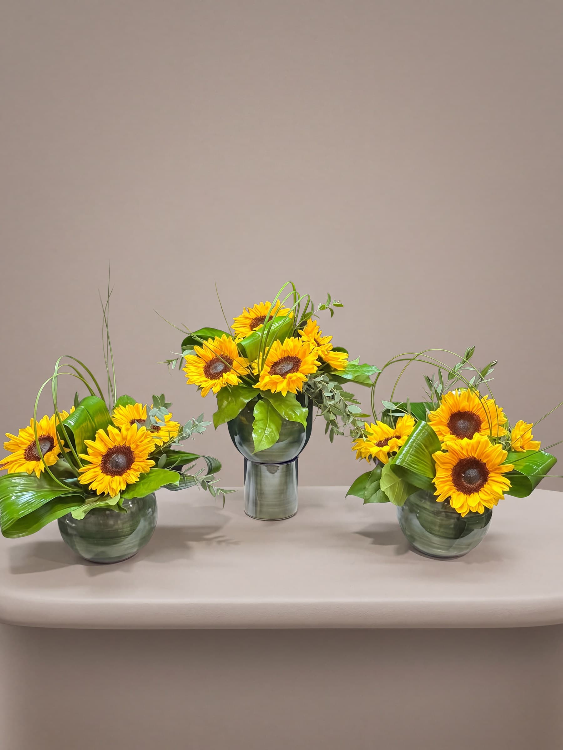 Set of three leaf-lined glass bubble bowls of sunflowers with eucalyptus and looped bear grass