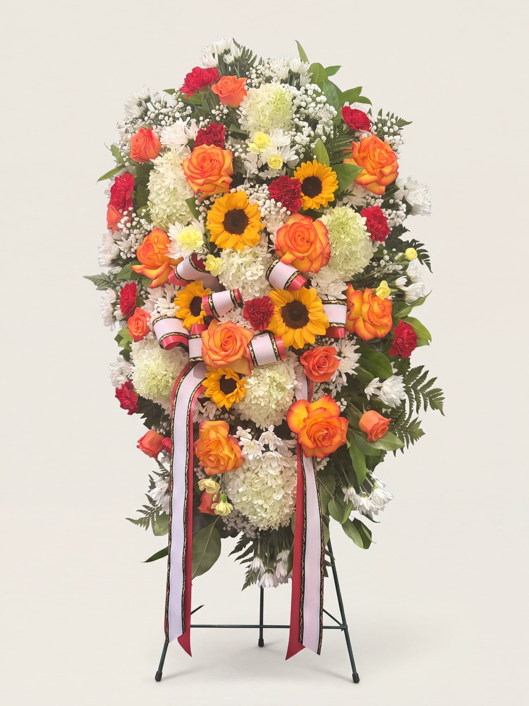 Standing funeral spray of sunflowers, orange roses, and white hydrangea with red carnations and a striped bow