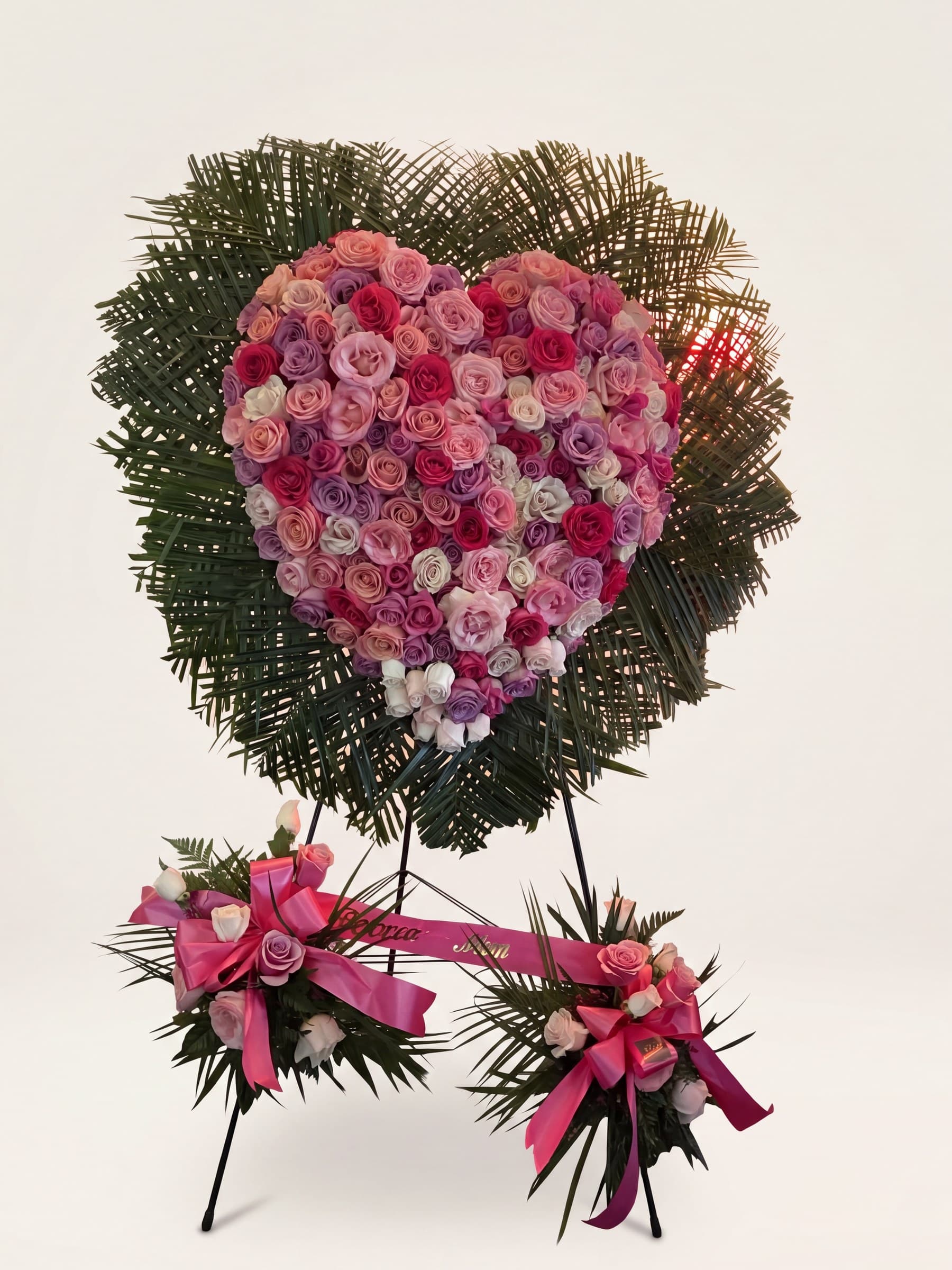 Solid funeral heart of pink and lavender roses on a fan of palm with pink bows on an easel