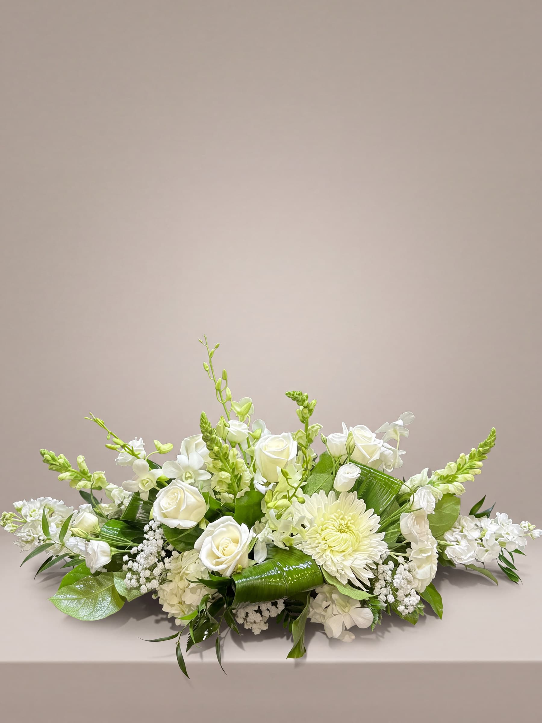 Long low all-white centerpiece of roses, dendrobium orchids, hydrangea, stock, snapdragon and a football mum over aspidistra leaves and fern