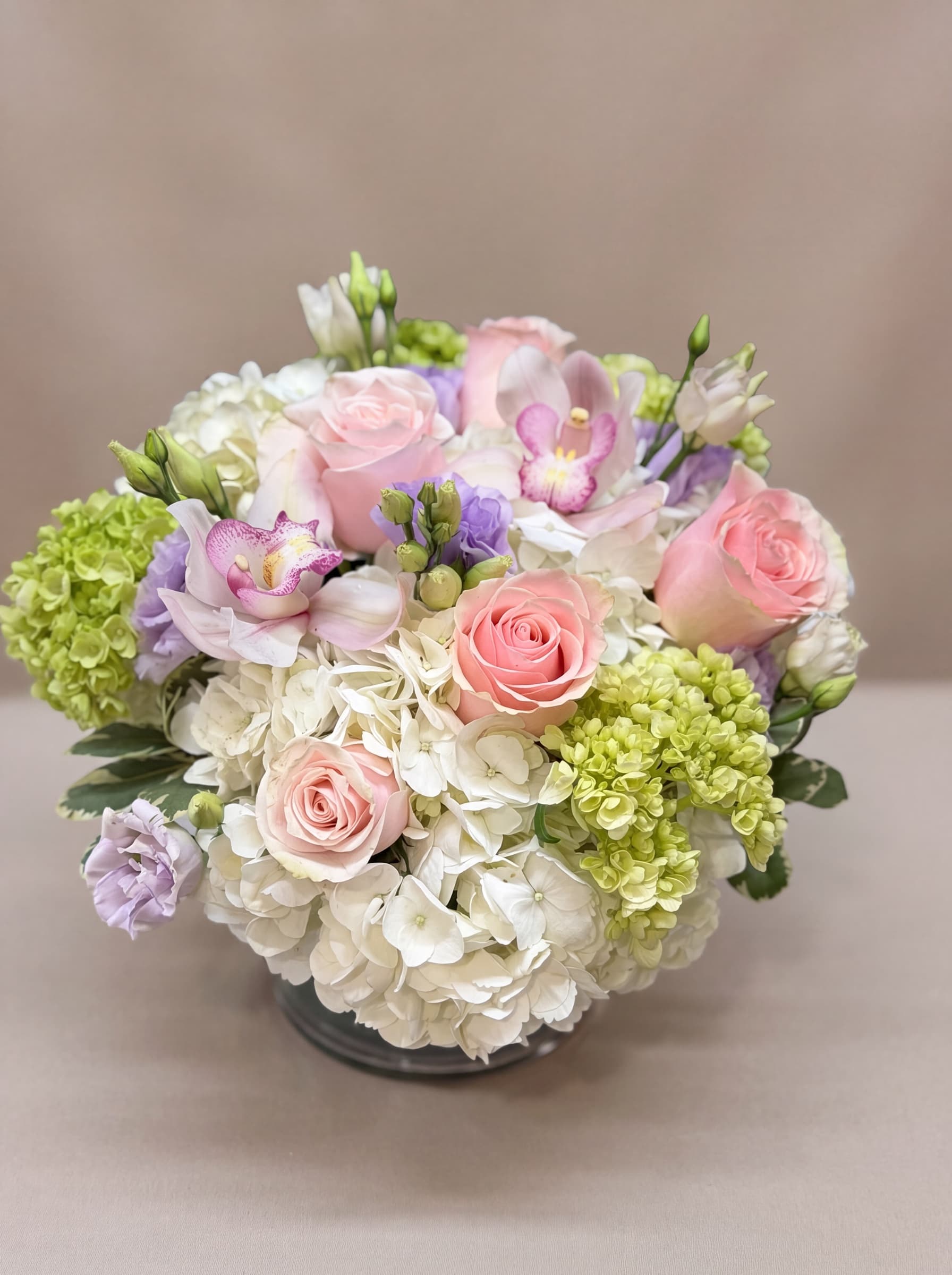Arreglo bajo y redondo de hortensias blancas, rosas blush, orquídeas cymbidium rosadas y lisianthus lavanda en cuenco de vidrio transparente