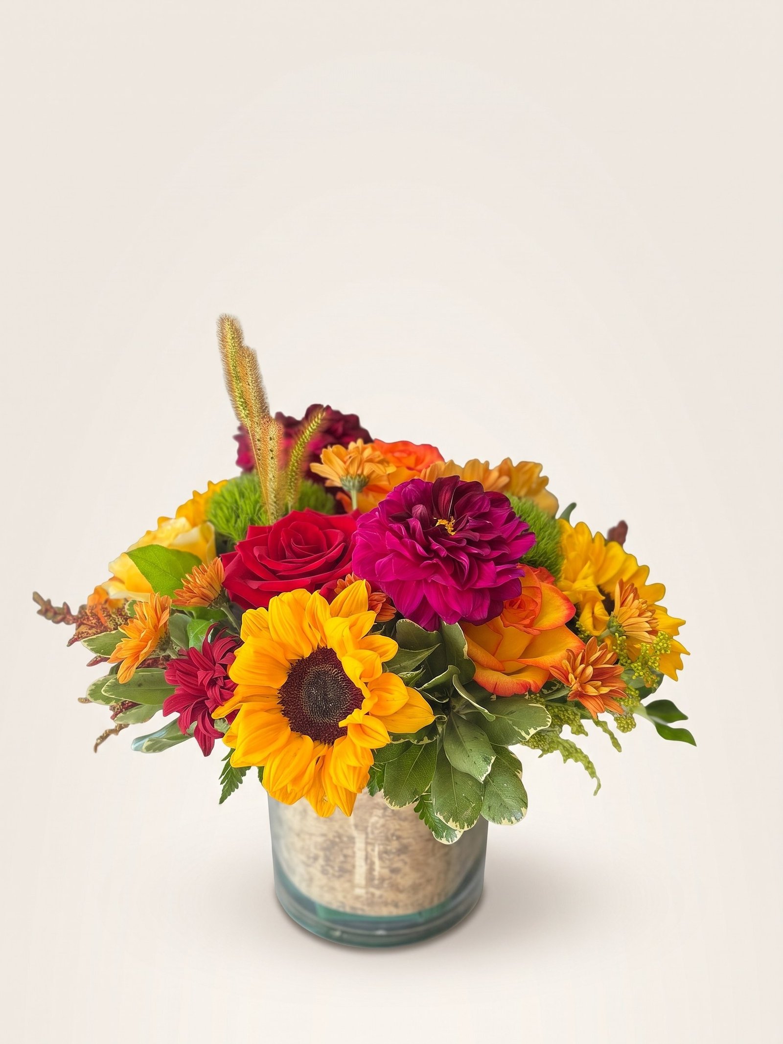 Fall arrangement of sunflowers, magenta dahlias, and red and orange roses in a glass cylinder