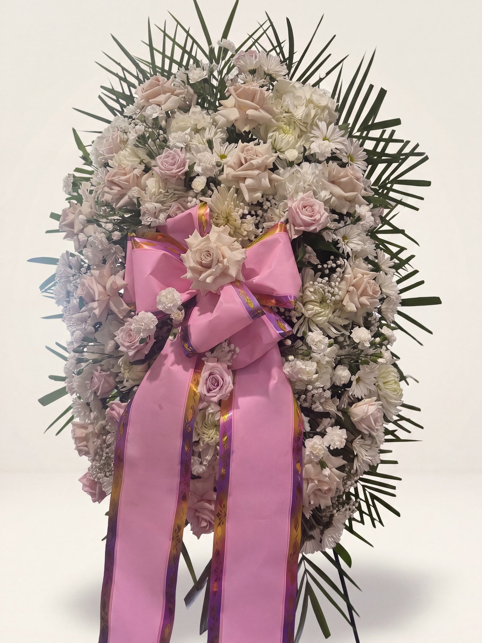 Oval standing funeral spray of cream and blush roses and white flowers framed in palm with a pink bow