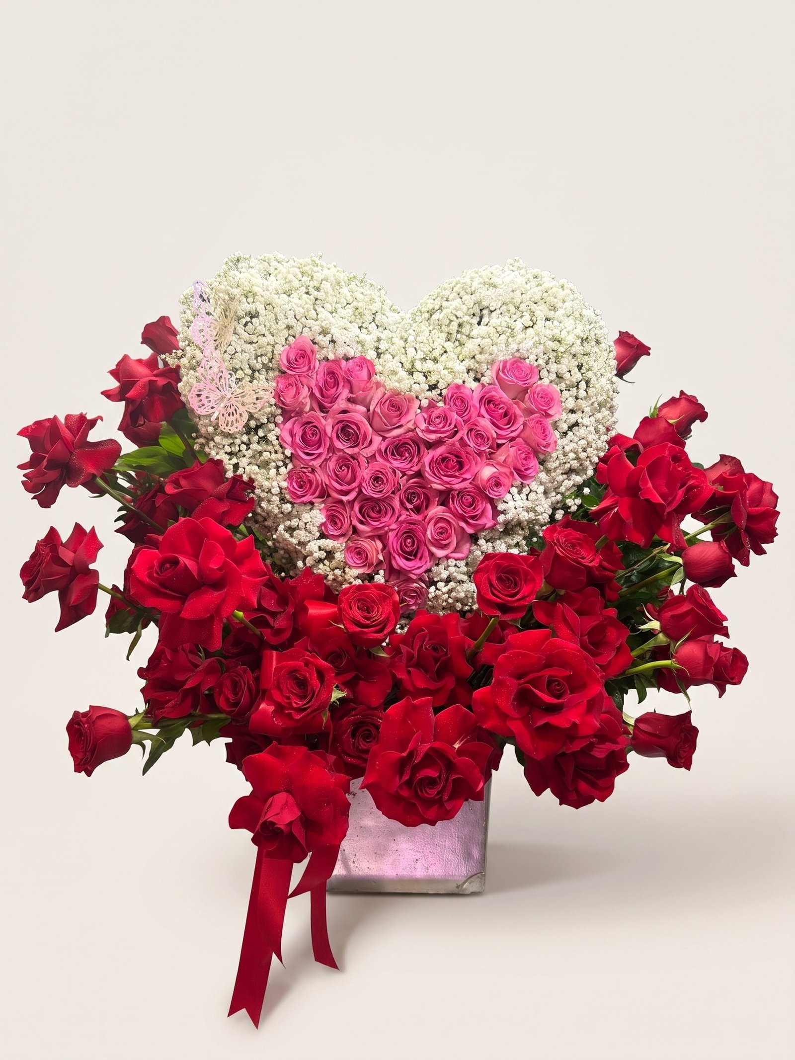 Heart of pink roses with a white baby's breath border over a mass of red roses, with a red satin bow in a silver cube, second view
