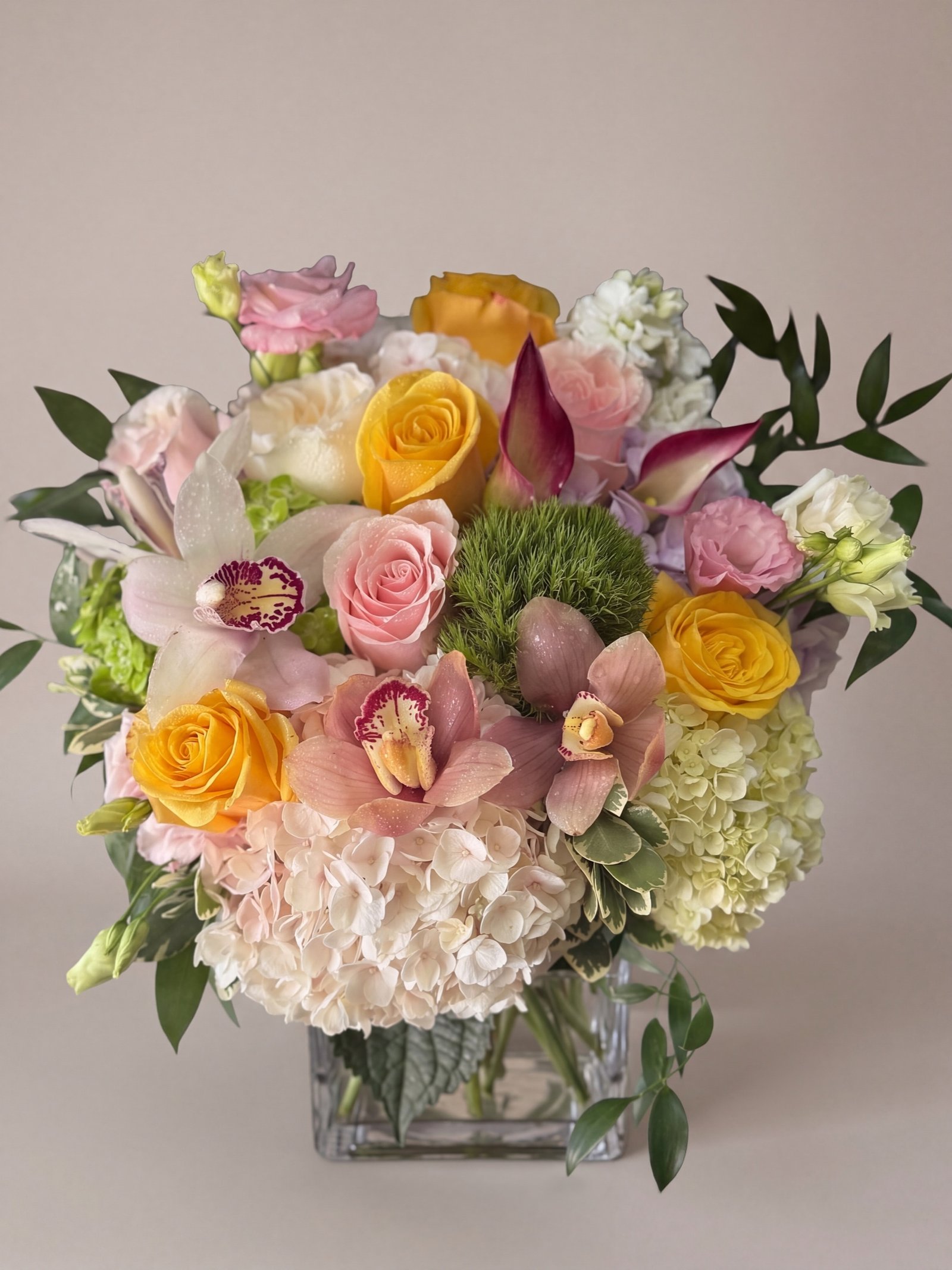 Arreglo pastel bajo de orquídeas cymbidium durazno, rosas amarillas y blush y hortensia crema en cubo de vidrio